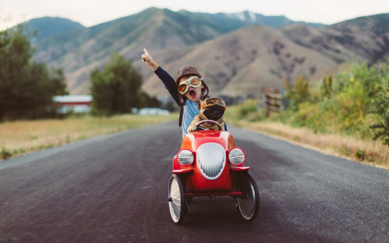 A young boy with flying goggles and flight cap races a red toy car with his pet and best friend French Bulldog along a small road in Utah, USA. Sometimes a road trip journey with your best friend and some fresh air in your face is the best medicine for the soul.