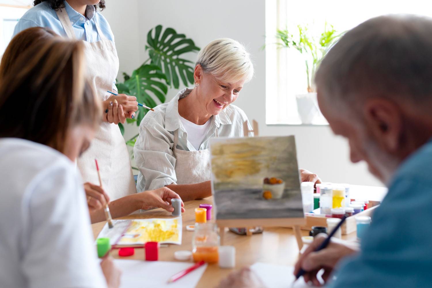 A group of old people engaged in a painting class, working with various colors and canvases on a bright, indoor table.